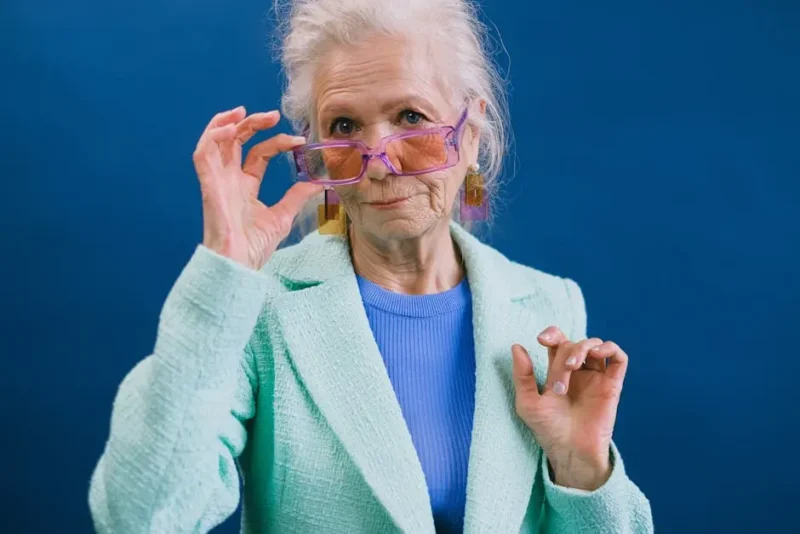 older woman posing for picture and holding onto her purple glasses on her face